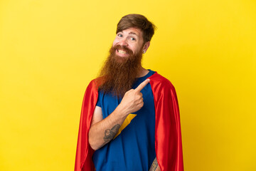 Redhead Super Hero man isolated on yellow background pointing to the side to present a product