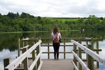 Young traveler with a backpack in lake dock enjoying the landscape. Young woman tourist watching...