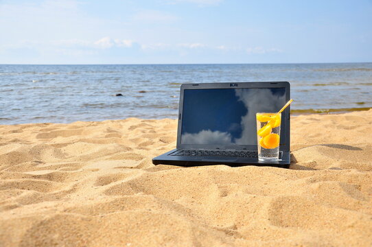 Remote work, a black laptop and a glass of fresh juice on a sandy beach.