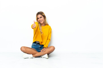 Young blonde Uruguayan woman sitting on the floor isolated on white background with thumbs up...