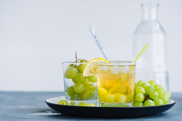 Refreshing summer drink in glass with ice cubes, green grape and lemon on the table.