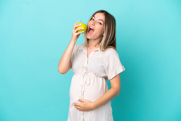 Young Romanian woman isolated on blue background pregnant and holding an apple and eating it