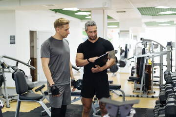 Fit man discussing workout plan with his trainer, personal coach before starting workout in the gym. Personal coach standing near young man and showing something on his clipboard