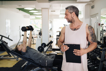 Muscular handsome trainer writing a fitness plan on clipboard for working out near his trainee in the fitness