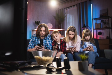 Portrait of caucasian parents with two kids sitting together on couch with personal smartphones in hands. Addiction from modern gadgets. Lifestyles concept.