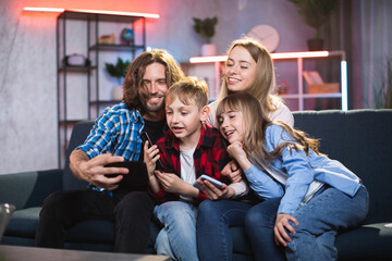 Fototapeta premium Positive caucasian family in casual wear taking selfie on smartphone while sitting on comfy couch. Young parents with little son and daughter having fun together at home.