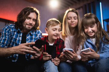 Positive caucasian family of four playing games on smartphones while sitting on comfy couch. Focus on modern gadgets in hands. Time spending of parents and kids.