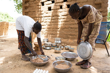 Two young black women preparing a meal for their family members in outdoor air kitchen in a west...