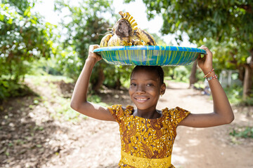 Pretty smiling black girl standing on a rural road in West Africa, carrying a plate full of bananas...