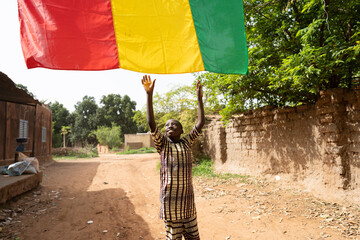 Cute little black African boy reaching out for the malian flag hanging over him on a rural village road