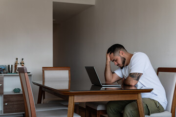young latin tattooed man in front of laptop stressed out and holding his head