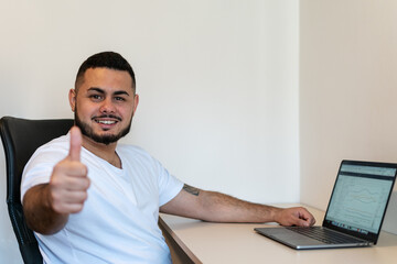 Young adult Latino young man looking at the camera while smiling and showing thumbs up. Next to a laptop computer