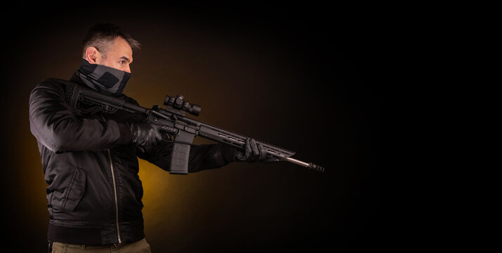 A man in dark clothes holds an automatic carbine in his hands. The face is covered with a mask, hands are in gloves. A figure with a weapon on a dark back - Powered by Adobe
