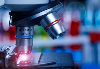 Close up shot of scientific microscope magnifying lens zooming monitoring on glass plate sample with red light flare in front of blurred red and blue test tubes background in hospital laboratory