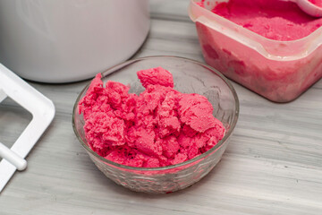 homemade cherry ice cream in a glass socket and a plastic container on the table next to an ice cream spoon and an ice cream maker