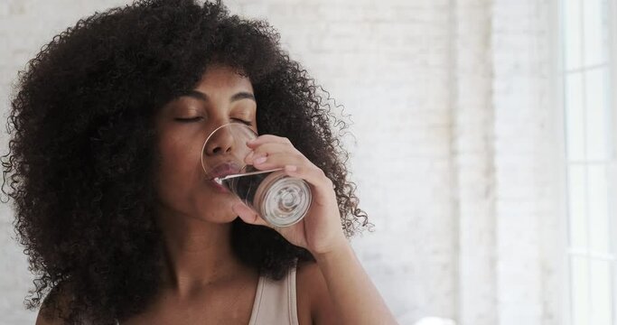 Young multiracial woman drinking water daily morning habit routine after morning exercises against large windows close view. Glass of still water healthy drink concept.