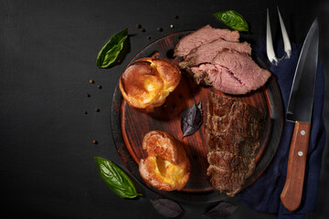 Roastbeef, yorkshire puddnings, basil leaves on a wooden cutting bords with napkin, fork and knife. Rustic style.