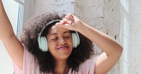 Happy hilarious black multiracial lady wearing headphones near the window listening to modern hip hop singing song with eyes closed resting in comfortable apartment at home
