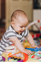 Cute chubby baby stands and plays with a children's music table. Lots of colorful toys everywhere.