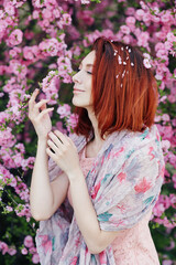 Naklejka premium Russian woman fashion close-up portrait. Beautiful sensual Caucasian young girl perfect skin creartive artistic makeup fashionable spring dress, short red hair. Girl standing in front of a sakura tree