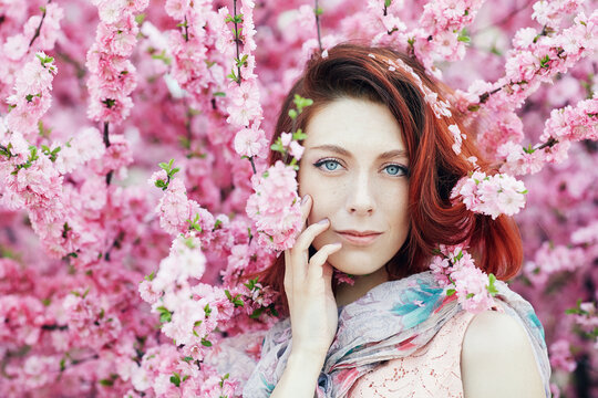 Close up portrait of a young and tender woman among beautiful pink flowers. Girl with bright blue eyes, clear skin, and long red hair