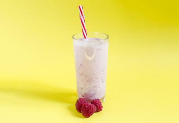 Milkshake in a transparent glass with raspberries and a red tube on a bright yellow background close-up. Cocktail of ice cream, milk and berries on a colored background