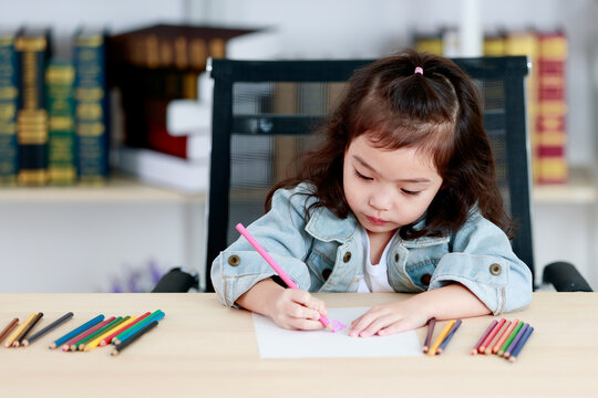 Little Asian girl concentrate to create picture drawing for school homework by pink pencil as art education while sitting on working desk of colored painting tools in reading room at home