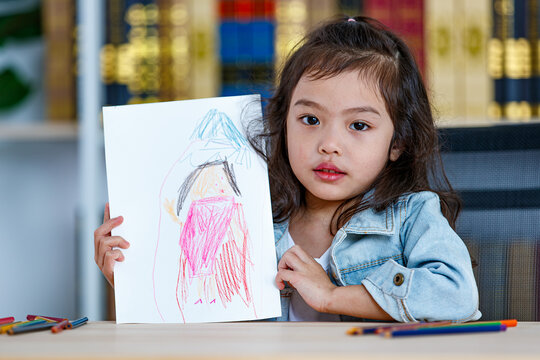 Lovely Asian girl happy and proud showing creative drawing of homework picture on white paper by colored pencil as art education while sitting on working desk of painting crayon at home
