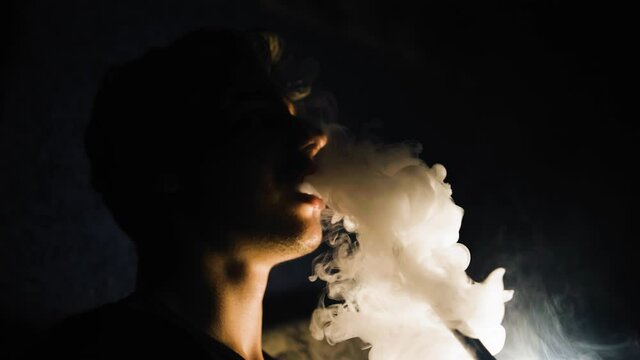 The guy smokes a hookah in a dark room and lets smoke out of his mouth. His silhouette is visible