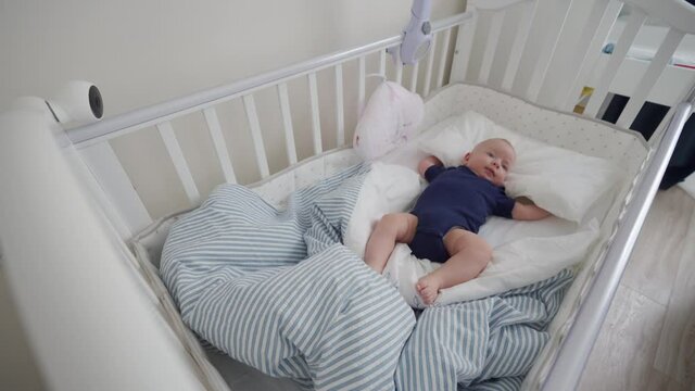 Baby wakes up in the crib, cute 3 months old baby boy lying in white baby cot, interior of a modern apartment bedroom. High quality 4k footage