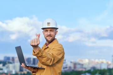 Bearded supervisor, foreman or construction inspector looking at camera and making Korean heart gesture. Contractor in hardhat and orange jacket holding computer laptop in front of sunny day blue sky