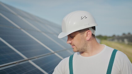 Portrait of happy male engineer in protective helmet looking to camera. Handsome man in uniform smiling while standing at solar power farm. Concept of green energy. Ecological future.