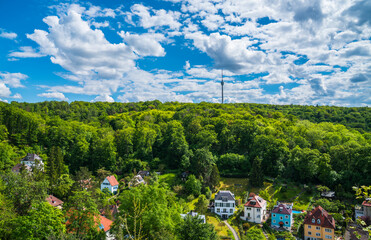 Germany, Stuttgart television tower building surrounded by green trees of forest on sunny day, aerial panorama drone view above the tree tops and houses