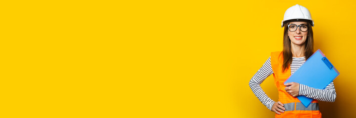 Young woman with a smile in a vest and hard hat holds a clipboard on a yellow background. Concept for construction, new building, renovation. Banner