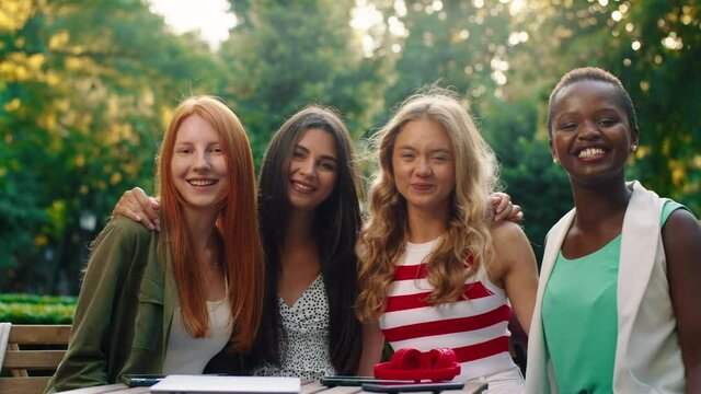 Four beautiful and charming women are sitting at a table with the trees in the background, they are hugging and smiling at each other, being very close