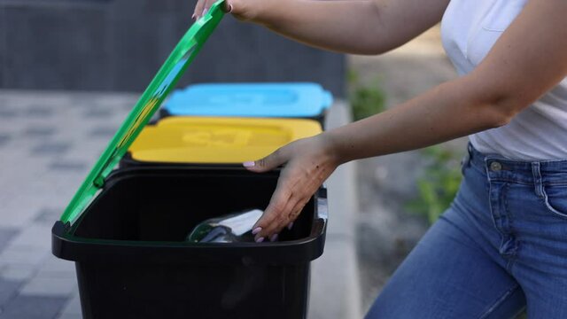 Close-up of woman's hand throwig empty glass bottle in recycling bin. Different colour of plastic bins in the park
