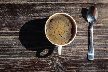 Cup of coffee in a white cup and a metal teaspoon on a gray-cinnamon wooden surface, light shadow,...