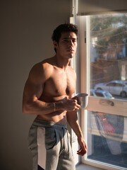 Young man shirtless next to window, holding a coffee cup