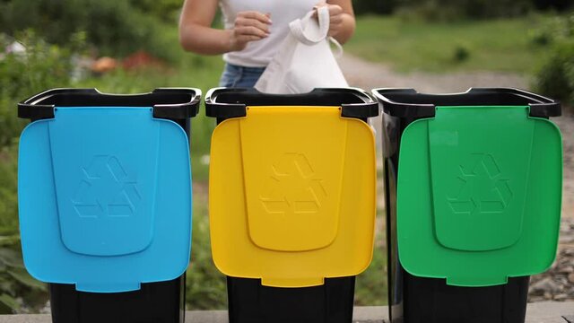 Middle secection of young woman taking from eco packager garbage and put them into recycling bins outddors. Concept of recycling. Tree bins, green, yeallow and blue. Lady put different kind of rabbish