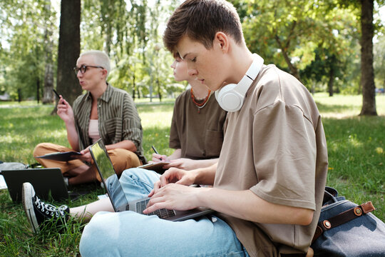 College student sitting on the grass and typing on laptop he sudying in the park together with other people