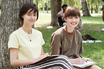 Portrait of two young women smiling at camera while sitting on the grass and studying outdoors in the park