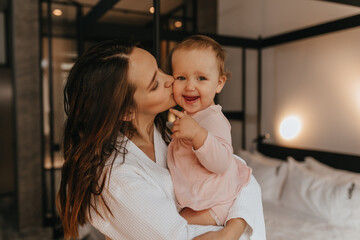 Mom kisses fair-haired child looking at camera with smile. Woman holds her daughter in her arms on background of white bed