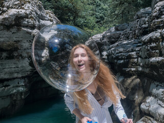 Laughing girl catches up with a shiny soap bubble looking through it against the backdrop of canyons and water