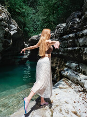 A longhaired girl stands in a swallow pose near a turquoise river in a rocky environment on a sunny day