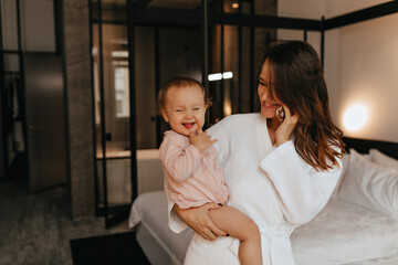 Lady in bathrobe in excellent mood is talking on phone, sitting on bed with perky female baby showing her tongue