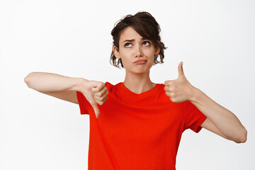Indecisive girl showing thumbs up and thumbs down, looking puzzled, cannot make choice, weighing...