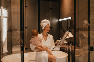 Charming woman after shower laughs, reading newspaper and holding baby. Portrait of mom and baby in bathroom