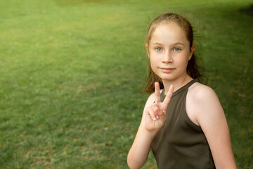 Young smiling teenager girl showing peace sign on green, outdoor park background