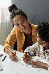 Young teacher sitting at desk with schoolboy they holding pencils and she teaching him to write