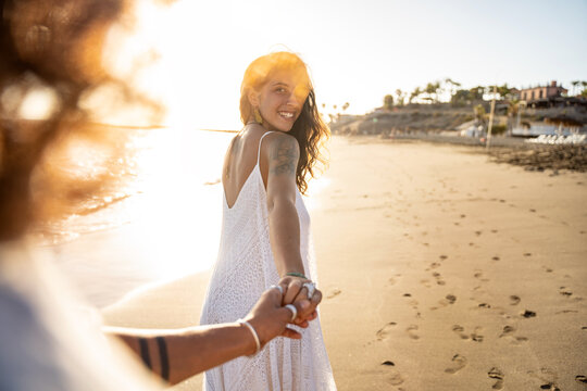 Lovely smiling beautiful woman holding hand of her boyfriend, looking at him, walking on the beach